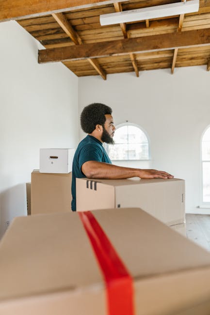 A young woman sitting on a wooden floor inside a room surrounded by numerous cardboard moving boxes of various sizes, some sealed with red tape and others labeled 'fragile.' She is wearing casual clot