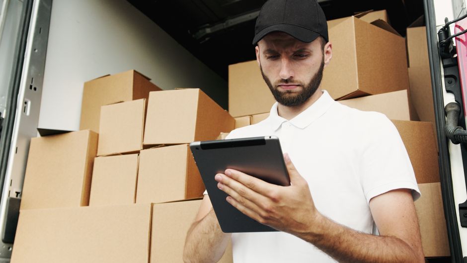 A male professional mover wearing a blue work uniform and a patterned bandana on his head is inside a property, standing beside open cardboard boxes filled with household items. He is holding a clipbo