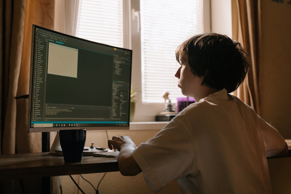 A person with short, light brown hair, wearing a light gray T-shirt, is seated at a wooden desk, engaged in packing or organising items for a house move. They sit on a gray desk chair in front of a la