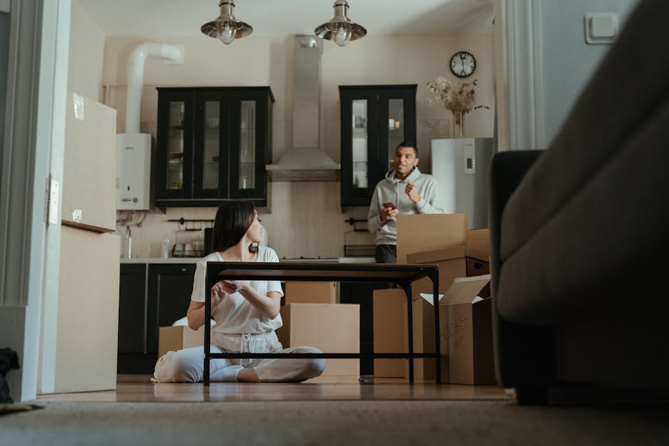 A woman with long blonde hair wearing a dark green t-shirt is inside a home, holding a clipboard and writing with a black marker. Behind her are several cardboard moving boxes, with one box open and p