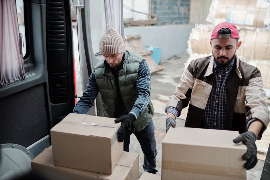 The image displays the rear of a white moving van with its double doors wide open, revealing an interior packed with various cardboard boxes, some of which are labeled 'fragile', and padded envelopes,